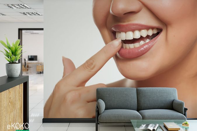 kobieta wskazuje palcem swoje białe zęby na jasnym tle - fototapeta do gabinetu stomatologicznego
